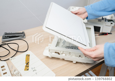 Hands carefully disassembling a flatbed scanner on a wooden desk, showing internal parts, cables and tools during repair and maintenance in a home office or small workplace setting. 139768802
