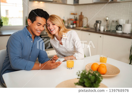 Happy couple laughing watching content on smartphone in kitchen Happy couple laughing watching content on smartphone in kitchen 139768976