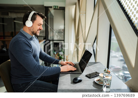 Adult man working remotely on laptop wearing headphones Adult man working remotely on laptop wearing headphones 139769689