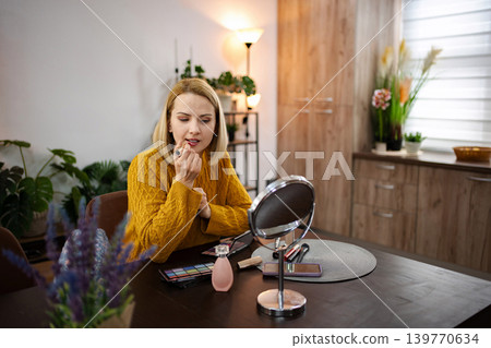 Creating an elegant look at home, a woman applies lipstick while seated at a well-lit vanity during early afternoon Creating an elegant look at home, a woman applies lipstick while seated at a well-lit vanity during early afternoon 139770634