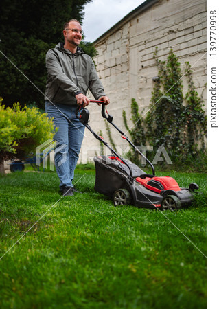Man mowing vibrant green lawn on a cloudy day in a residential garden Man mowing vibrant green lawn on a cloudy day in a residential garden 139770998