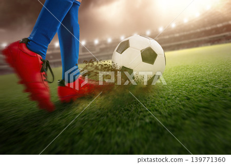 Dynamic action shot of a soccer player in red cleats kicking a ball on a green field under stadium lights with flying dirt and motion blur 139771360