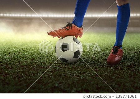 Detailed view of a soccer player standing with one foot on a soccer ball on a green pitch in a brightly lit stadium at night Detailed view of a soccer player standing with one foot on a soccer ball on a green pitch in a brightly lit stadium at night 139771361
