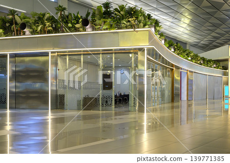 A wide-angle view of a modern airport terminal interior featuring a sleek glass storefront, decorative indoor plants, and a polished reflecting floor under high architectural ceilings A wide-angle view of a modern airport terminal interior featuring a sleek glass storefront, decorative indoor plants, and a polished reflecting floor under high architectural ceilings 139771385