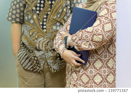 Two colleagues in traditional Indonesian batik attire standing in an office environment. One woman wearing a hijab is holding a blue document folder, representing a professional meeting 139771387