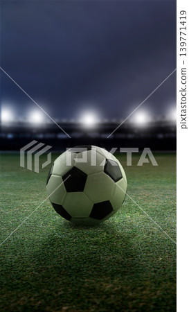 A classic black and white soccer ball sits on a green grass field under bright stadium floodlights at night, creating a dramatic athletic atmosphere A classic black and white soccer ball sits on a green grass field under bright stadium floodlights at night, creating a dramatic athletic atmosphere 139771419