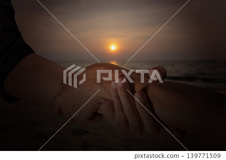 A close-up shot showing two hands clasped in a supportive gesture against the backdrop of a beautiful sunset over the calm ocean waves at dusk A close-up shot showing two hands clasped in a supportive gesture against the backdrop of a beautiful sunset over the calm ocean waves at dusk 139771509