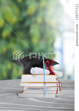 A black graduation mortarboard with a blue tassel sits atop a stack of books and a diploma tied with a red ribbon, set against a blurred green background 139771511