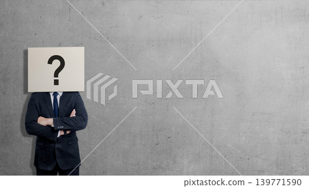 A businessman in a suit stands with arms crossed against a gray concrete wall, his head obscured by a sign featuring a large black question mark, symbolizing anonymity, doubt, or curiosity A businessman in a suit stands with arms crossed against a gray concrete wall, his head obscured by a sign featuring a large black question mark, symbolizing anonymity, doubt, or curiosity 139771590