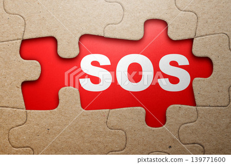The word SOS is revealed through a missing section of a cardboard jigsaw puzzle, set against a red background to signify a distress call or critical need The word SOS is revealed through a missing section of a cardboard jigsaw puzzle, set against a red background to signify a distress call or critical need 139771600