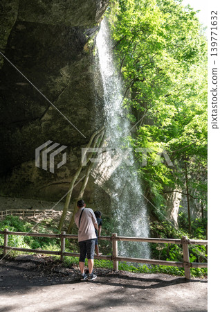 A parent and child enjoy the waterfall behind the waterfall, with the water splashing. 139771632