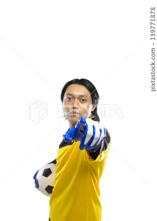 Portrait of a young Indonesian southeast asian male soccer goalkeeper in a yellow jersey and gloves, holding a soccer ball and pointing forward against a white background 139771878