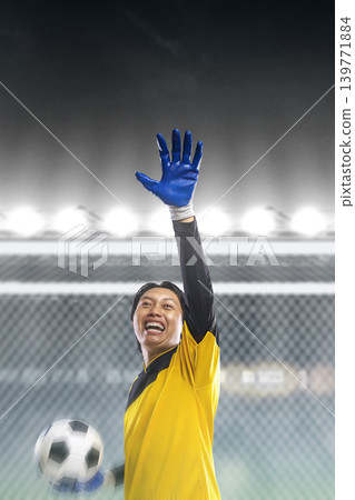 Professional Indonesian southeast asian soccer goalie in a yellow jersey and blue gloves celebrates a save, smiling and holding a ball under bright stadium lights at night during a match Professional Indonesian southeast asian soccer goalie in a yellow jersey and blue gloves celebrates a save, smiling and holding a ball under bright stadium lights at night during a match 139771884