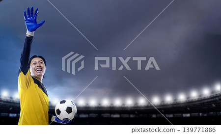 A happy Indonesian southeast asian male soccer goalkeeper in a yellow jersey, wearing blue gloves, holds a football and raises one arm in victory at a lit stadium under a dark sky A happy Indonesian southeast asian male soccer goalkeeper in a yellow jersey, wearing blue gloves, holds a football and raises one arm in victory at a lit stadium under a dark sky 139771886