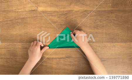 A child's hands folding green origami paper into a triangle, the moment the corners meet. A child's hands folding green origami paper into a triangle, the moment the corners meet. 139771890