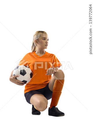 Full length portrait of a young Indonesian southeast asian woman in orange football uniform kneeling and holding a soccer ball, isolated against a plain white background 139772064