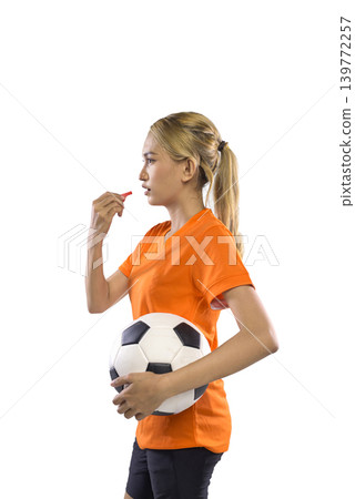 A young Indonesian southeast asian woman in an orange jersey and black shorts holds a soccer ball and blows a whistle against a white background A young Indonesian southeast asian woman in an orange jersey and black shorts holds a soccer ball and blows a whistle against a white background 139772257
