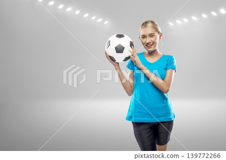 A joyful young Indonesian southeast asian woman with a bright smile holds a soccer ball against a blurred stadium background with glowing lights. Perfect for sports and fitness themes A joyful young Indonesian southeast asian woman with a bright smile holds a soccer ball against a blurred stadium background with glowing lights. Perfect for sports and fitness themes 139772266