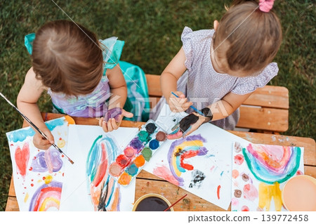 Two little girls painting rainbows at a wooden table in the garden Two little girls painting rainbows at a wooden table in the garden 139772458