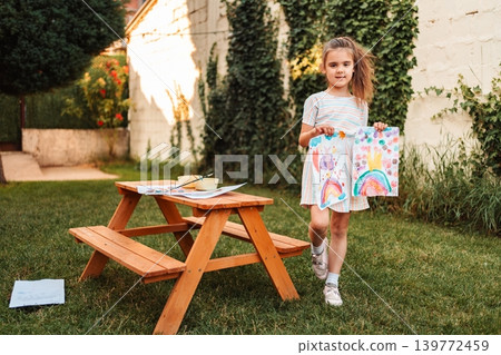 Little girl proudly showing her drawings in the garden Little girl proudly showing her drawings in the garden 139772459