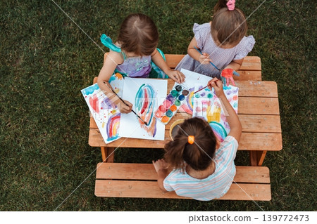Three little girls painting with watercolors at a wooden table in a garden 139772473
