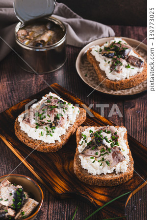 Slices of black bread with ricotta and sardines canned in oil on a board on a table vertical view 139773025