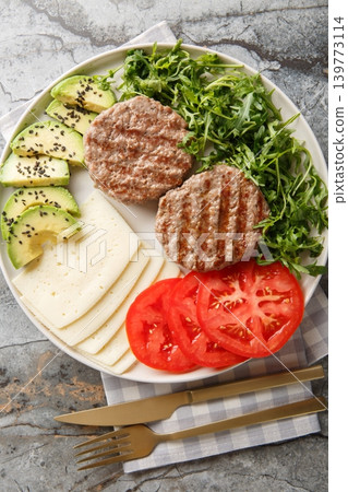 Diet beef burger with cheese, avocado, grilled patty, tomatoes and arugula close-up on a plate. Vertical top view Diet beef burger with cheese, avocado, grilled patty, tomatoes and arugula close-up on a plate. Vertical top view 139773114