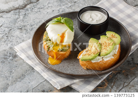 Close-up of two toasts with poached egg and basil pesto, avocado, and cream cheese served with sour cream on a plate. Horizontal Close-up of two toasts with poached egg and basil pesto, avocado, and cream cheese served with sour cream on a plate. Horizontal 139773125