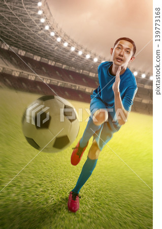 Young Indonesian southeast asian soccer player in blue jersey running on a professional stadium grass field with a ball in motion. High energy action shot with bright stadium lights Young Indonesian southeast asian soccer player in blue jersey running on a professional stadium grass field with a ball in motion. High energy action shot with bright stadium lights 139773168