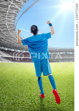 Rear view of a Indonesian southeast asian soccer player in blue uniform celebrating a win with arms raised in a sunny stadium on a green grass field Rear view of a Indonesian southeast asian soccer player in blue uniform celebrating a win with arms raised in a sunny stadium on a green grass field 139773169