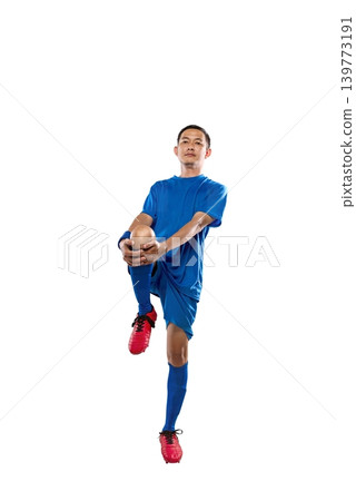 Professional Indonesian southeast asian soccer player in blue uniform performing a knee-to-chest leg stretch, isolated on a white background. Ideal for fitness and sports marketing Professional Indonesian southeast asian soccer player in blue uniform performing a knee-to-chest leg stretch, isolated on a white background. Ideal for fitness and sports marketing 139773191