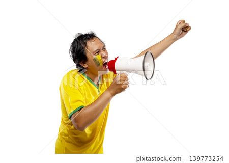 An excited Indonesian southeast asian male soccer fan wearing a yellow and green jersey shouting into a megaphone and cheering with his fist raised, isolated on white background An excited Indonesian southeast asian male soccer fan wearing a yellow and green jersey shouting into a megaphone and cheering with his fist raised, isolated on white background 139773254