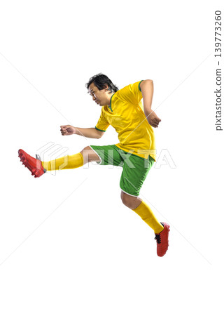 Full length shot of a professional Indonesian southeast asian male soccer player in a yellow and green uniform performing a dynamic jumping kick, isolated on white background. 139773260