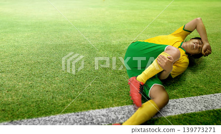 A Indonesian southeast asian male athlete in a yellow and green soccer uniform lies on the grass field, clutching her knee and grimacing in pain after suffering a sports injury during a match A Indonesian southeast asian male athlete in a yellow and green soccer uniform lies on the grass field, clutching her knee and grimacing in pain after suffering a sports injury during a match 139773271