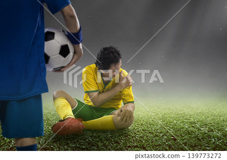 A Indonesian southeast asian soccer player sits on the grass pitch in pain, holding his injured shoulder while another player stands nearby holding a soccer ball during a match under stadium lights A Indonesian southeast asian soccer player sits on the grass pitch in pain, holding his injured shoulder while another player stands nearby holding a soccer ball during a match under stadium lights 139773272