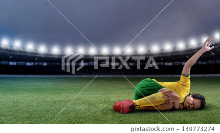 Professional Indonesian southeast asian male soccer player in yellow and green uniform lies on the stadium grass in pain, clutching her injured knee during a match under bright floodlights 139773274