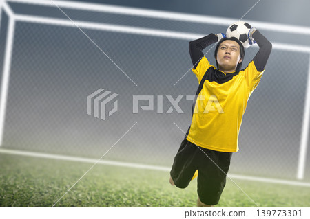 Indonesian southeast asian professional soccer goalkeeper in a yellow jersey jumps to catch the ball during a match on a grass field with a goalpost in the background 139773301