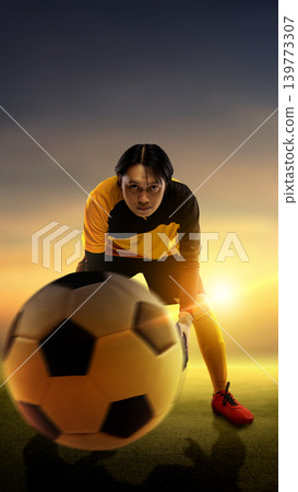 A focused Indonesian southeast asian male soccer goalkeeper in a black and yellow kit ready to catch a ball on a green field during a beautiful sunset. Motion blur on the football adds dynamic energy 139773307