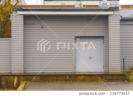 Gray metal shutter with security door. Commercial building entrance featuring metal gate and safety sign Gray metal shutter with security door. Commercial building entrance featuring metal gate and safety sign 139773657