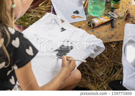 Child paints cat design on white t-shirt during craft workshop with fabric paint, brush, art supplies. Concept of school holidays, kids activity, textile art, handmade fashion, hobby Child paints cat design on white t-shirt during craft workshop with fabric paint, brush, art supplies. Concept of school holidays, kids activity, textile art, handmade fashion, hobby 139773800
