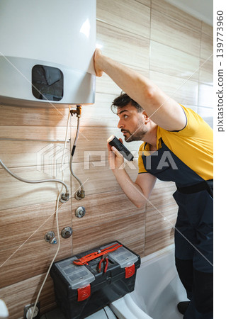Plumber inspecting water heater in bathroom using flashlight 139773960