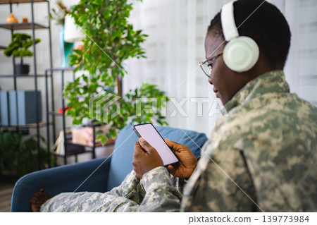 Soldier relaxing on sofa using smartphone and listening music with headphones Soldier relaxing on sofa using smartphone and listening music with headphones 139773984