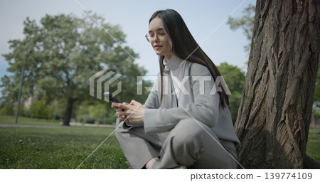 The shot is handheld and shows a young female student in casual clothes wearing glasses sitting on the grass under a tree in a park and using a mobile phone. The shot is handheld and shows a young female student in casual clothes wearing glasses sitting on the grass under a tree in a park and using a mobile phone. 139774109