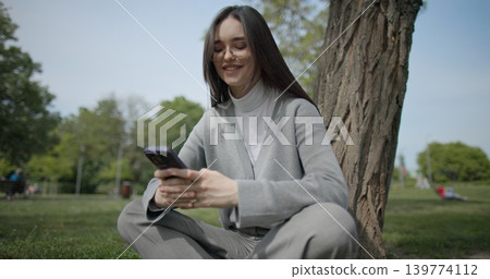 The shot is handheld and shows a young female student in casual clothes wearing glasses sitting on the grass under a tree in a park and using a mobile phone. The shot is handheld and shows a young female student in casual clothes wearing glasses sitting on the grass under a tree in a park and using a mobile phone. 139774112