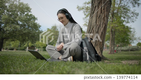 A hand-held shot of a young beautiful female student in casual clothes, wearing glasses and headphones, sitting on the grass under a tree in a city park and using a laptop while typing on the keyboard 139774114