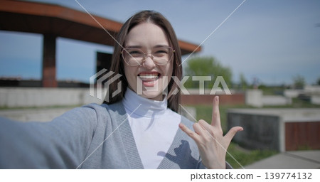 The shot is handheld and shows a young female student in casual clothes wearing glasses, sitting and recording a selfie video on her phone, holding it in her hand and gesturing with her other hand. 139774132