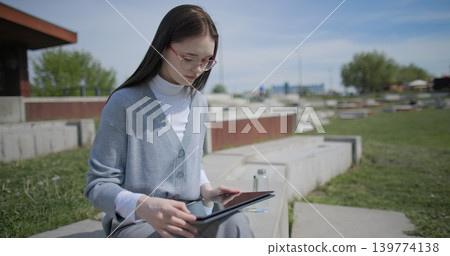 The shot is handheld and shows a young female student in casual clothes wearing glasses sitting on a concrete bench in an urban park and working on a tablet. The shot is handheld and shows a young female student in casual clothes wearing glasses sitting on a concrete bench in an urban park and working on a tablet. 139774138