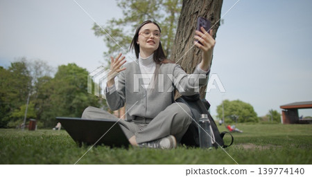 A hand-held shot of a young, beautiful female student in casual clothes, wearing glasses and headphones, sitting on the grass under a tree in a city park, using a laptop and talking on a mobile phone. A hand-held shot of a young, beautiful female student in casual clothes, wearing glasses and headphones, sitting on the grass under a tree in a city park, using a laptop and talking on a mobile phone. 139774140