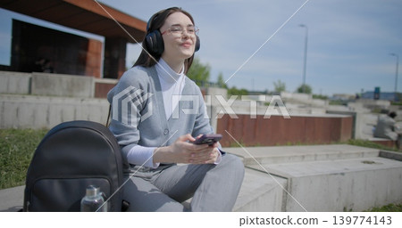 A hand-held shot of a young female student in casual clothes, wearing glasses and headphones, sitting on a concrete in an urban park on a sunny summer day and listening to music from a mobile phone A hand-held shot of a young female student in casual clothes, wearing glasses and headphones, sitting on a concrete in an urban park on a sunny summer day and listening to music from a mobile phone 139774143