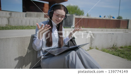 A hand-held shot of a young female student in casual clothes, glasses, and headphones making a video call on a laptop while sitting on a concrete bench in an urban park. 139774149